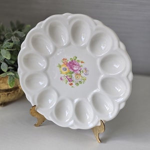 May include: A white ceramic egg plate with a scalloped edge and twelve oval indentations arranged in a circle. The center of the plate features a colorful floral decal with pink, yellow, and purple flowers surrounded by green leaves.