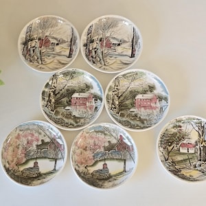 May include: A set of seven round ceramic plates featuring detailed landscape illustrations. Each plate displays a different scene, including red barns, stone walls, trees, and water features, rendered in a palette of green, brown, red, and white.