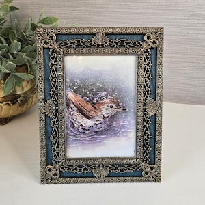May include: A rectangular picture frame with a dark blue inner border and an ornate, metallic bronze-colored filigree outer edge. The frame contains an illustration of a brown and white bird splashing in water with visible droplets.