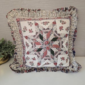 May include: A square throw pillow featuring a central star quilt pattern in shades of pink, green, and white with floral motifs. The pillow is bordered by a ruffled edge made of cream-colored lace and a floral fabric trim, creating a decorative finish.