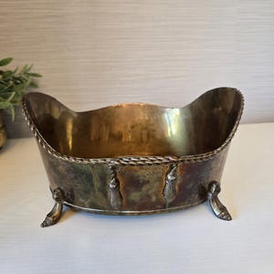 May include: An oval-shaped brass bowl with a tarnished finish, featuring a decorative rope-style rim, hanging tassel accents along the exterior, and four curved feet. The interior is smooth and reflective, while the exterior shows patina.