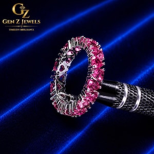 Könnte beinhalten: Ein silberner Eternity-Ring mit einer durchgehenden Reihe leuchtend pinker, herzförmiger Edelsteine. Die Metallfassung zeigt eine Reihe kleiner herzförmiger Aussparungen entlang des Seitenprofils. Das Logo GEN Z JEWELS ist oben links zu sehen.