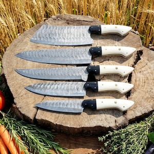 Könnte beinhalten: Ein Set aus fünf Küchenmessern mit strukturierten Stahlklingen und cremefarbenen Griffen mit schwarzen Akzenten. Die Messer sind auf einer Holzoberfläche angeordnet und zeigen ihre unterschiedlichen Größen und Formen für verschiedene kulinarische Aufgaben.