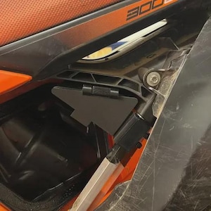 Può includere: Primo piano di un componente nero di una moto con un coperchio incernierato. Il componente è montato su un telaio di moto arancione e nero. L'immagine mostra i dettagli del design e della costruzione della moto.