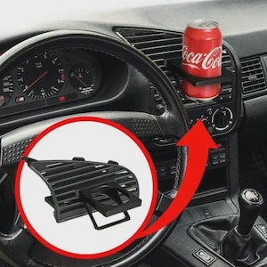 Puede incluir: Una taza de cerámica blanca con asa negra y el texto "But First, Coffee" en negrita negra. La taza se encuentra sobre un fondo borroso, sugiriendo un ambiente acogedor.