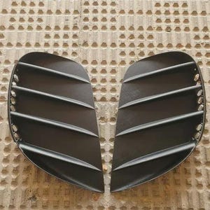 以下が含まれることがあります： 水平スラットを備えた、2つの黒い半円形の自動車用エアベント。 ベントはマットな素材でできており、テクスチャ加工された明るい色の背景に対して並んで配置されています。 各ベントには、一連の平行な隆起したバーがあります。