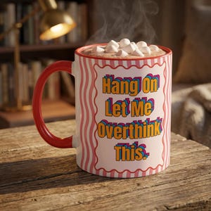 May include: A white ceramic mug with a red handle and red wavy vertical stripes. The front features a scalloped rectangular frame containing the orange text "Hang On Let Me Overthink This." The mug is filled with a hot beverage topped with marshmallows.