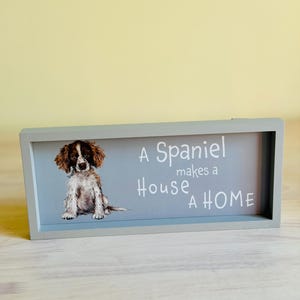 May include: A rectangular wooden sign with a light gray frame. The sign features a watercolor-style image of a spaniel dog and the text "A Spaniel makes a House a HOME" in white lettering against a light blue background.