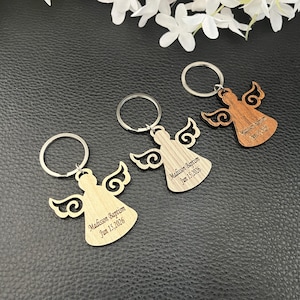 May include: Three wooden angel keyrings with silver key rings. Each angel has detailed wings and is engraved with the text "Madison Baptism Jun 15, 2026". One keyring is light wood, one is medium wood, and one is dark wood.