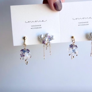 Könnte beinhalten: Handgefertigte Ohrringe mit kleinen, durchscheinenden lila und blauen Blüten, goldfarbenem Metall und baumelnden tropfenförmigen Anhängern. Die Ohrringe werden auf einer weißen Karte mit dem Text "HANDMADE ACCESSORY" präsentiert.