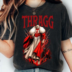 May include: Dark gray t-shirt featuring the word "THRAGG" in red, above a superhero illustration. The superhero is in a red and white costume with a fur collar, red cape, and lightning effects.