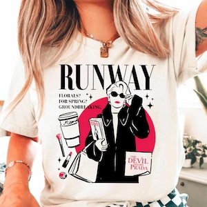 May include: White t-shirt with a graphic design featuring the word "RUNWAY" above an illustration of a woman in sunglasses and a black coat. The design includes text, a coffee cup, and a book.
