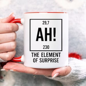 May include: White ceramic mug with a red interior and handle. The mug features a black square with the text "29,7 AH! 230 THE ELEMENT OF SURPRISE" in a bold, sans-serif font. The mug is held in a person's hands.