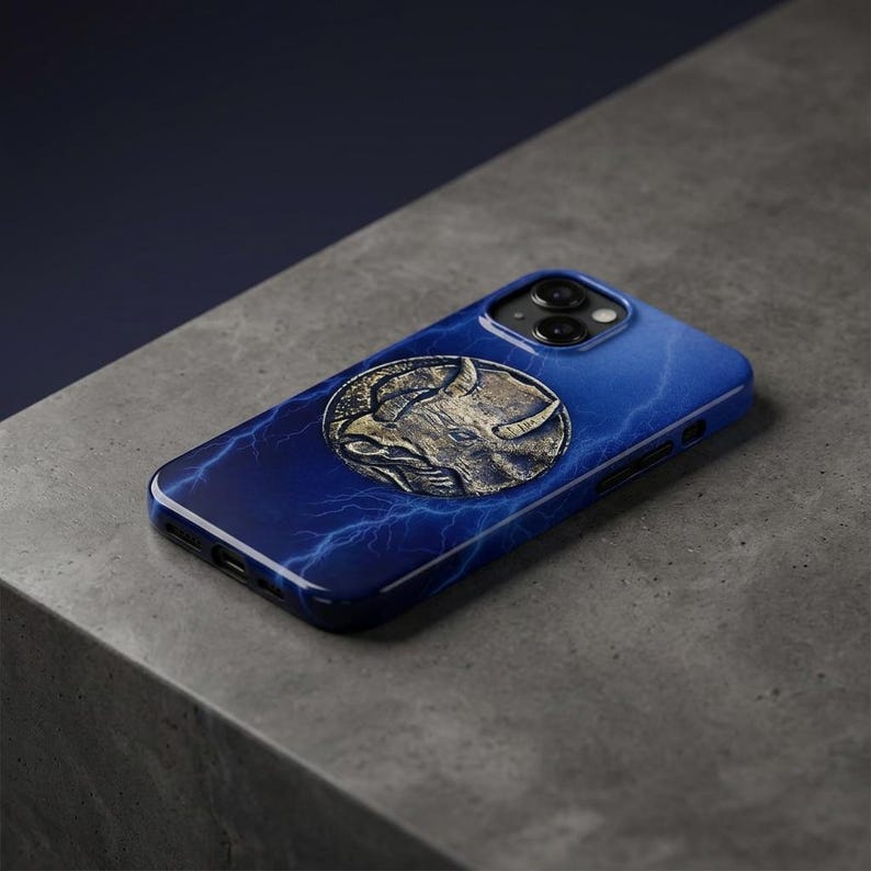 May include: A blue iPhone case with a metallic gold emblem of a horned animal. The case has a lightning bolt design and a glossy finish. The phone case is on a gray surface.