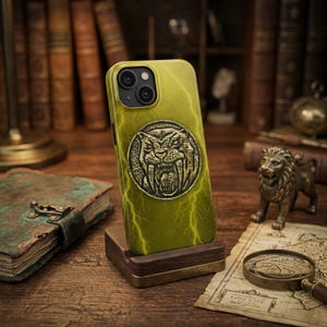 May include: A yellow phone case with a saber-toothed tiger emblem and lightning bolt design. The case is displayed on a wooden stand, showcasing its intricate details and unique style. The case is designed for a smartphone.