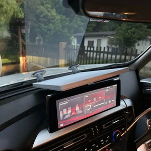 Könnte beinhalten: Ein Armaturenbrett im Auto mit einem großen, rechteckigen Digitaldisplay, das eine Navigationsschnittstelle mit roten und weißen Grafiken anzeigt. Über dem Display ist ein graues Regal montiert.
