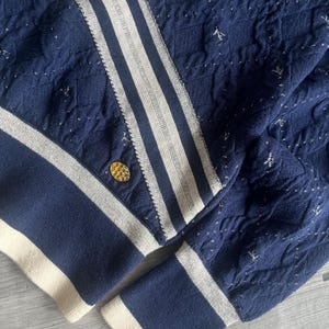 May include: Navy blue knit jacket with white stripes and accents. The jacket features a gold button and a textured pattern with silver flecks. The cuffs and hem are trimmed with a cream-colored band.
