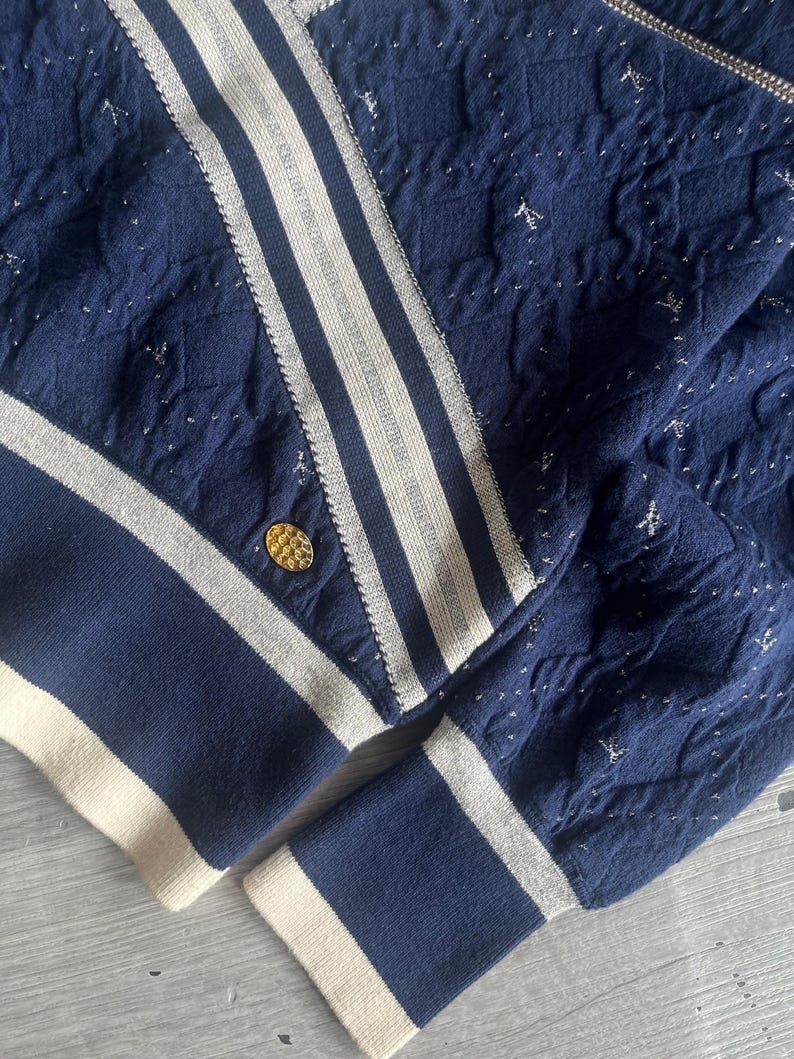 May include: Navy blue knit jacket with white stripes and accents. The jacket features a gold button and a textured pattern with silver flecks. The cuffs and hem are trimmed with a cream-colored band.