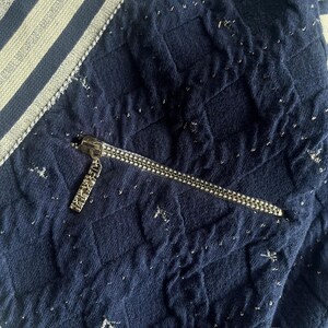 May include: Close-up of a navy blue quilted fabric with silver flecks. A silver zipper is partially open, revealing a pocket. The fabric has a textured appearance and is accented with a white and silver striped trim.