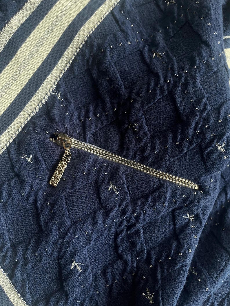 May include: Close-up of a navy blue quilted fabric with silver flecks. A silver zipper is partially open, revealing a pocket. The fabric has a textured appearance and is accented with a white and silver striped trim.
