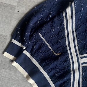 May include: Navy blue jacket with white stripes and a zipper. The jacket has a textured pattern with small white accents. The cuffs and collar are navy blue with white trim. The jacket has a gold zipper pull.
