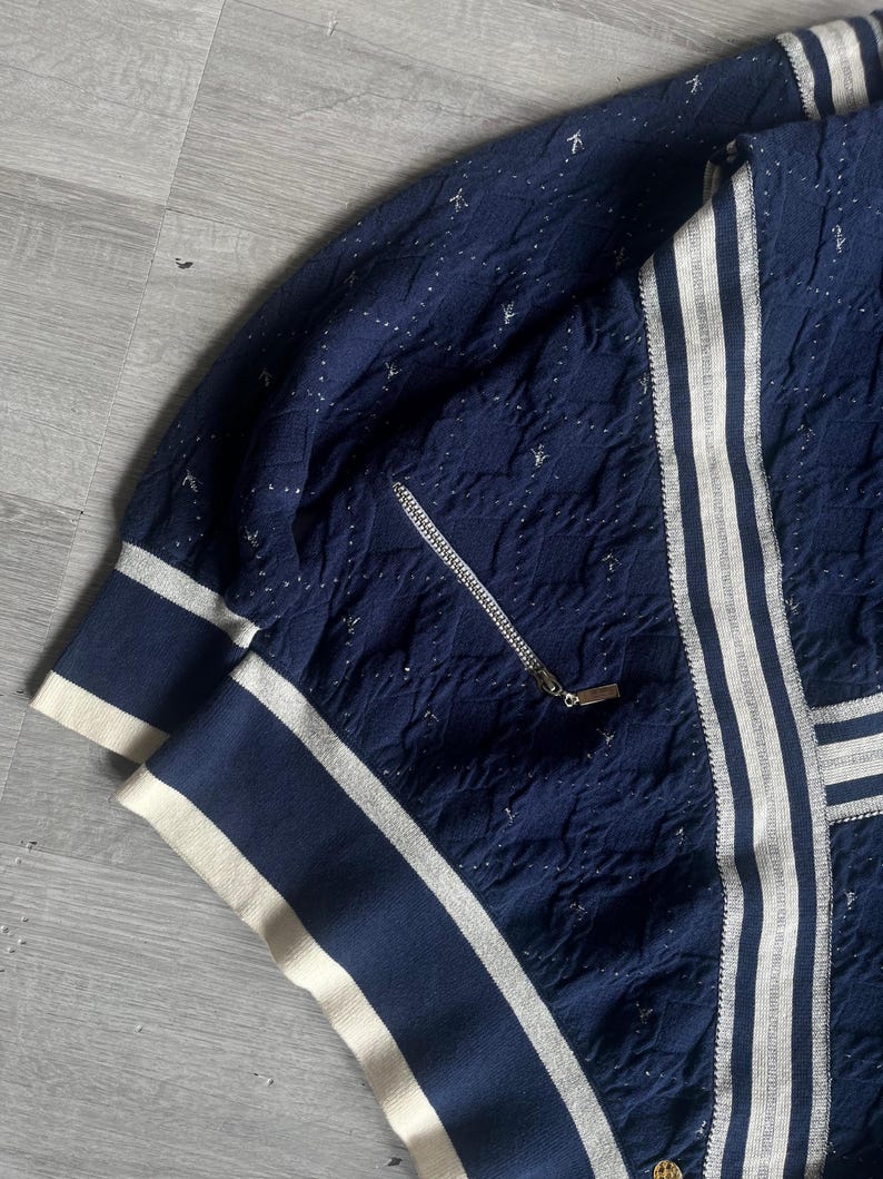 May include: Navy blue jacket with white stripes and a zipper. The jacket has a textured pattern with small white accents. The cuffs and collar are navy blue with white trim. The jacket has a gold zipper pull.