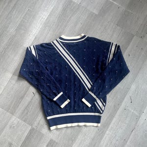 May include: Navy blue sweater with white stripes and trim. The sweater features a textured knit pattern and a crew neck. The sleeves and hem have white and navy blue bands. A diagonal white stripe runs across the front.