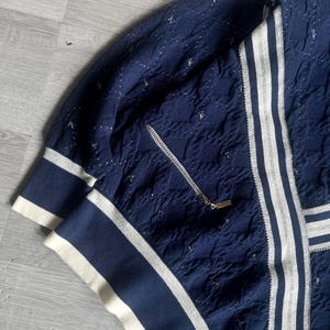 May include: Navy blue zip-up jacket with white stripes and accents. The jacket features a textured pattern and a silver zipper. The cuffs and collar have a white and navy blue striped design. The jacket is made of a soft, comfortable material.