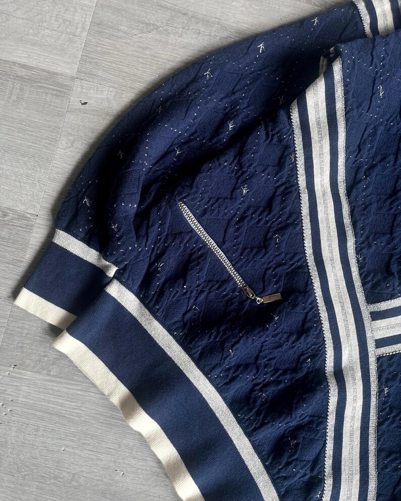 May include: Navy blue zip-up jacket with white stripes and accents. The jacket features a textured pattern and a silver zipper. The cuffs and collar have a white and navy blue striped design. The jacket is made of a soft, comfortable material.