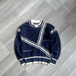 May include: Navy blue sweater with white stripes and detailing. The sweater has a textured knit pattern, ribbed hem, and two small zipped pockets. It features a white collar and cuffs, and a gold emblem near the hem.