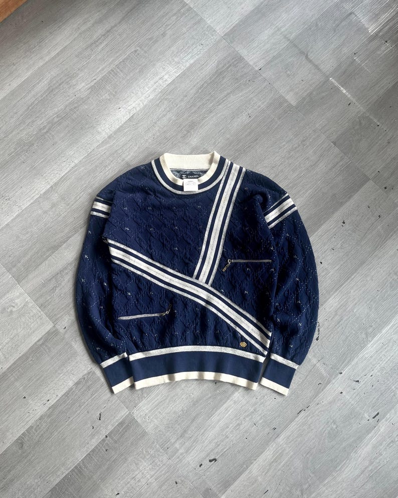May include: Navy blue sweater with white stripes and accents. The sweater features a textured knit pattern, a ribbed hem, and two small zippered pockets. The design includes a white collar and cuffs, and a gold emblem near the hem.
