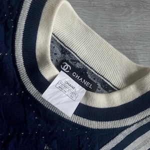 May include: Close-up of a navy blue sweater with a cream-colored collar and silver stripes. The sweater has a textured pattern and a tag that reads "CHANEL". The tag also includes the style number and size 40.