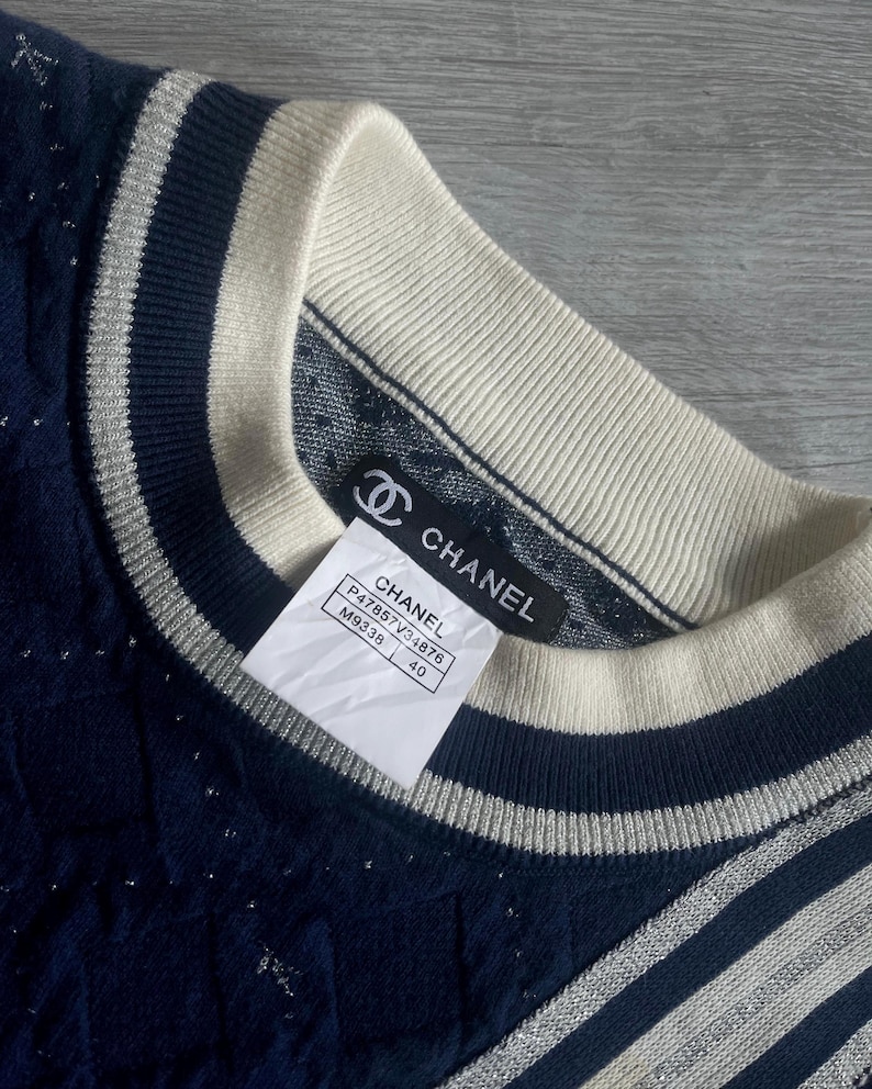 May include: Close-up of a navy blue sweater with a cream-colored collar and silver stripes. The sweater has a textured pattern and a tag that reads "CHANEL". The tag also includes the style number and size 40.