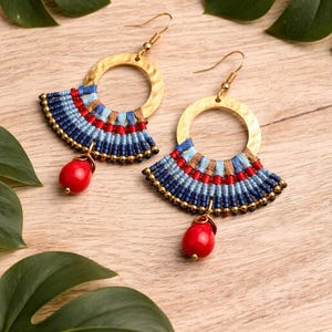 Puede incluir: Un par de pendientes hechos a mano con una parte superior circular dorada y un diseño en forma de abanico. El abanico presenta hilos tejidos azules, rojos y marrones, con una cuenta roja en forma de lágrima colgando debajo. Los pendientes están sobre una superficie de madera.