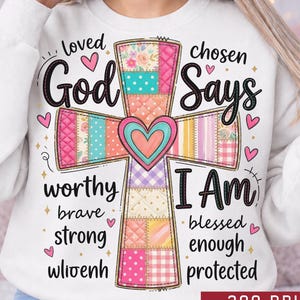 May include: White sweatshirt with a colorful cross design. The cross is made of patchwork patterns in pink, blue, and yellow. Text includes "God Says I Am Loved Chosen" and other inspirational words. Hearts and stars accent the design.