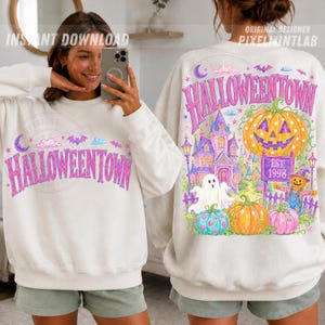 May include: A cream-colored long-sleeve sweatshirt featuring a graphic print with the text HALLOWEENTOWN. The design includes a purple haunted house, a large orange jack-o'-lantern, a white ghost, and smaller pumpkins in pink and orange.