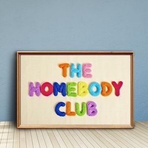 Könnte beinhalten: Ein rechteckiges gerahmtes Wandbild mit dem Schriftzug THE HOMEBODY CLUB, der aus dreidimensionalen, bunten Strickgarnbuchstaben besteht. Die Buchstaben sind in drei Reihen auf einem hellbeigen, strukturierten Stoffhintergrund angeordnet.