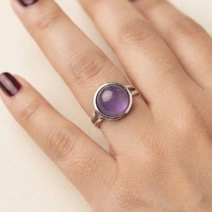 Op de afbeelding: Een zilveren ring met een gladde, ronde, in cabochon geslepen paarse amethist in een eenvoudige metalen zetting. De ringband is gespleten in twee dunne, parallelle zilveren draden die samenkomen bij de basis van de zetting van de steen.