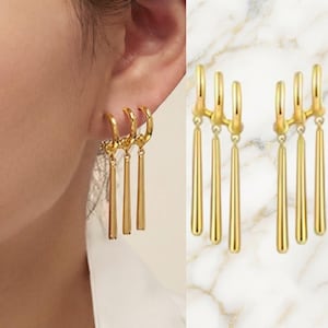 May include: A pair of gold-toned earrings featuring three curved hoops connected to three dangling, slender, cylindrical bars. The design creates a layered effect with the bars hanging at varying lengths, providing a polished metallic finish.