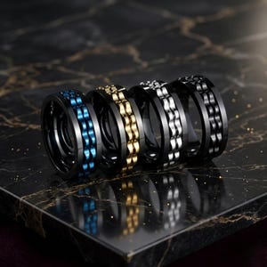 May include: Four black rings with contrasting centre bands in blue, gold, silver, and black. The rings are presented on a dark, marble-effect surface. Each ring features a distinctive central design, offering a modern and stylish aesthetic.