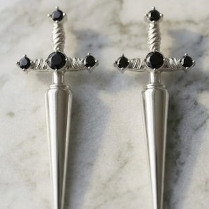 May include: A pair of silver-toned dagger-shaped earrings. Each earring features a pointed blade design and a cross-shaped top adorned with black gemstones. The earrings are displayed on a marble surface.