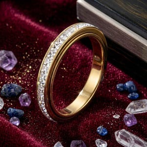 May include: A gold ring with a band of clear, sparkling gemstones. The ring is set against a deep red velvet surface, with scattered gemstones in shades of purple, blue, and clear. The ring's design is elegant and eye-catching.