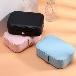 May include: Three rectangular jewelry boxes in black, pink, and light blue. Each box has a rounded top and a clasp closure with a silver-colored button. The boxes are stacked on a light orange surface.