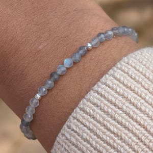 May include: A delicate bracelet featuring small, round, light blue beads strung together. The bracelet has a silver accent bead and is worn on a wrist. The wrist is covered by a cream-colored sweater.