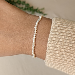 May include: A fine bracelet made of small, round, off-white pearls. The bracelet is strung together and worn on a wrist, with a beige sweater sleeve in the background. The pearls are uniform, creating a classic and elegant appearance.