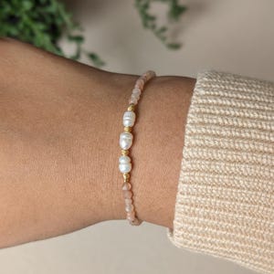 May include: A delicate bracelet featuring small, light pink beads interspersed with three large, white pearls. The pearls are separated by gold-coloured metal accents. The bracelet is worn on a wrist, with a neutral-coloured jumper in the background.