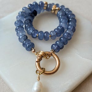 Pode incluir: Uma pulseira de fio duplo com contas azuis de textura rachada, espa&ccedil;adores de metal dourado e um grande fecho de mola circular dourado. Um &uacute;nico pingente de p&eacute;rola branca em forma de l&aacute;grima pende do fecho, adicionando um detalhe distinto.