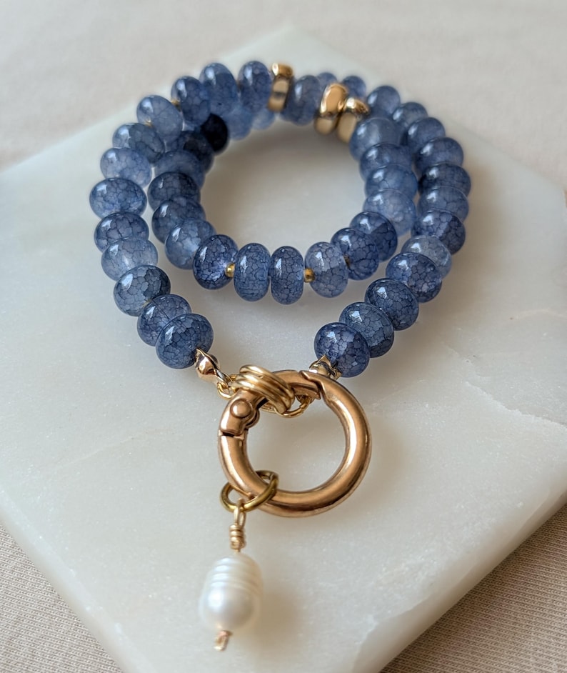 Pode incluir: Uma pulseira de fio duplo com contas azuis de textura rachada, espa&ccedil;adores de metal dourado e um grande fecho de mola circular dourado. Um &uacute;nico pingente de p&eacute;rola branca em forma de l&aacute;grima pende do fecho, adicionando um detalhe distinto.