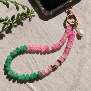 May include: A phone charm strap featuring a strand of round, faceted beads in shades of green and pink. The strap includes gold-toned metal accents, a gold-colored circular clasp, and a single white pearl-like bead dangling from the attachment point.