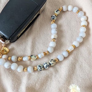 May include: A beaded keychain with white and dalmatian jasper beads, gold accents, and a gold clasp. The keychain is next to a black car key.